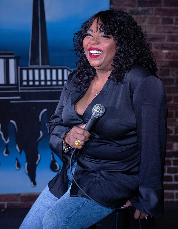 Cocoa Brown sitting on a stool on stage at a comedy club and holding a microphone
